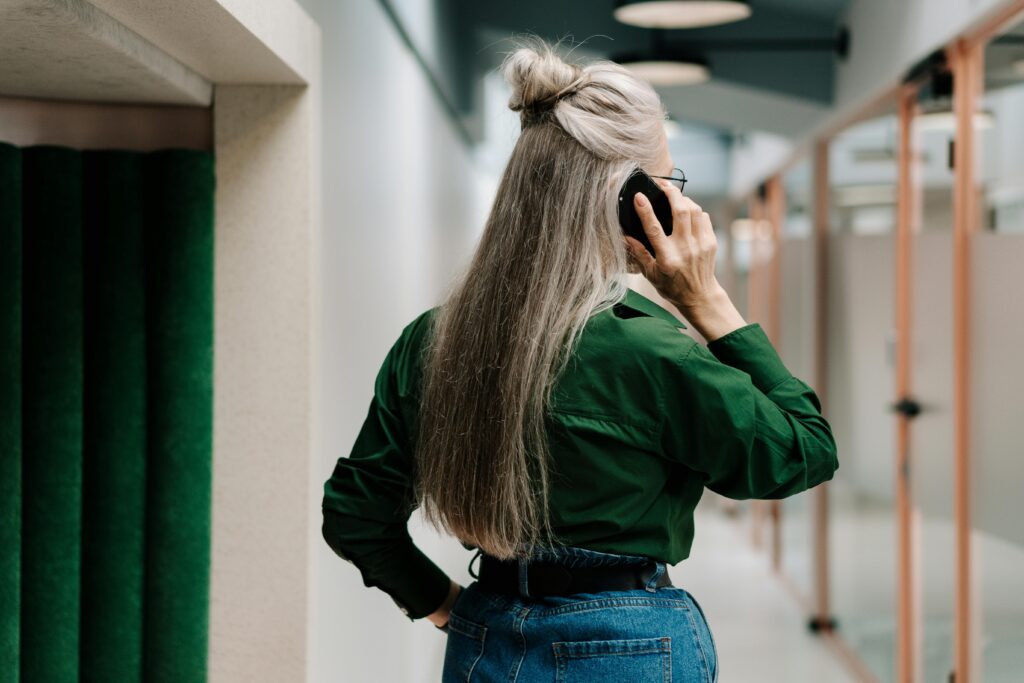 Woman with long grey hair in a half-up bun, seen from behind while talking on a phone in an office corridor, wearing a green jacket and jeans.