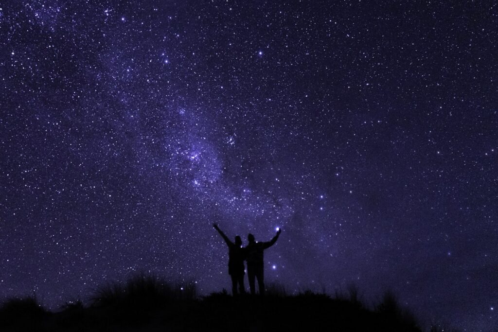 The silhouettes of two people standing side by side on a dark hilltop, both with arms raised toward a vivid deep-blue night sky filled with stars and a bright band of the Milky Way.