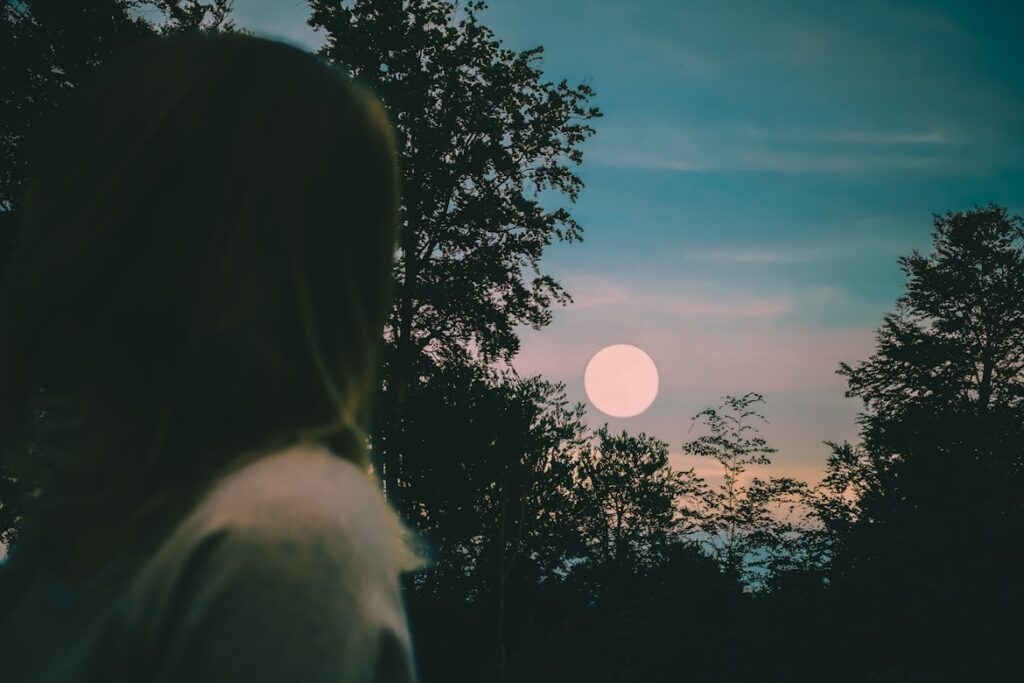 A person seen from behind, silhouetted against a dusky blue and orange sky, watching a bright full moon through the dark outlines of surrounding trees.