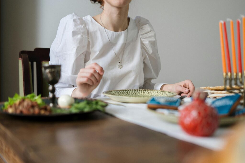 A woman in a white blouse seated at a wooden table set for a Passover Seder, with a Seder plate of herbs and egg in the foreground, matzah, a goblet, a pomegranate, and a menorah with pink candles.