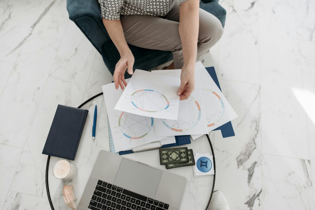 Overhead view of a person sitting cross-legged on a white marble floor, holding printed astrological birth charts with colorful wheel diagrams, surrounded by a laptop, navy notebooks, a lit candle, and a coaster showing the Gemini zodiac symbol.