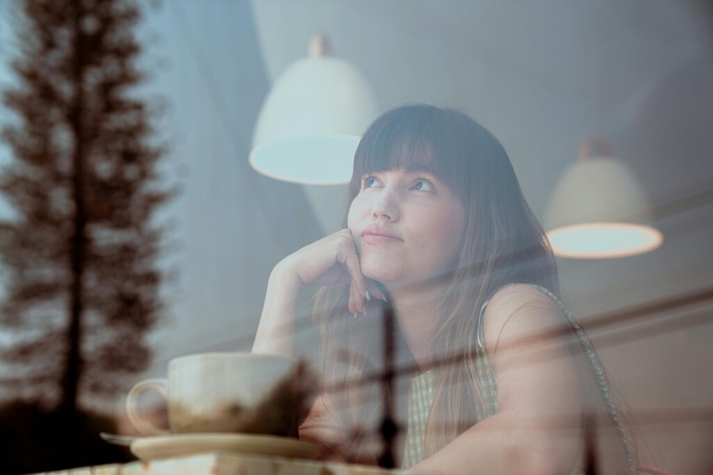 Young woman with long brown hair and bangs gazes thoughtfully upward while sitting in a cafe. Her chin rests on her hand, a white coffee cup before her. Pendant lamps hang overhead and trees reflect in the window glass.