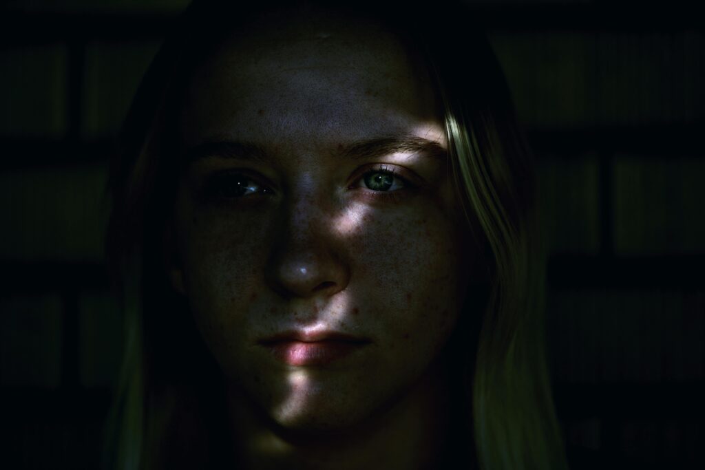 Dramatic low-key portrait of a young woman's face in darkness. A narrow strip of light illuminates only her eyes and the bridge of her nose, revealing freckles and light blonde hair.