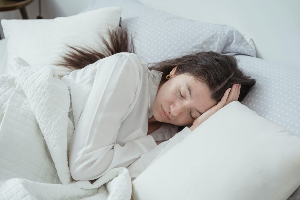 Woman with brown hair sleeps peacefully on her side in white bedding, her head resting on a pillow, eyes closed, wearing a white long-sleeved top.