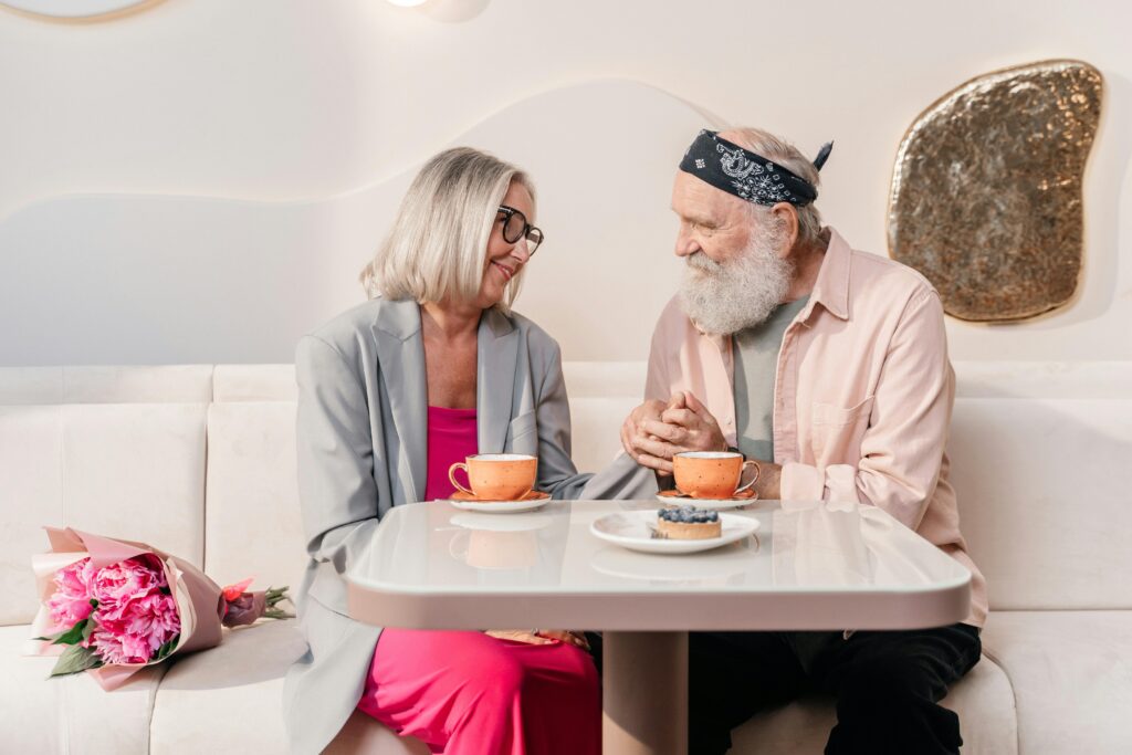 An older couple sitting close together at a small white table in a bright café, holding hands between two orange coffee cups, with a bouquet of pink peonies resting on the table beside them. Both are smiling and looking at each other.