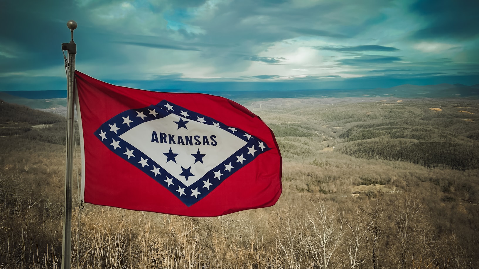 Arkansas state flag with red field and blue diamond containing white stars waving against a rural landscape.