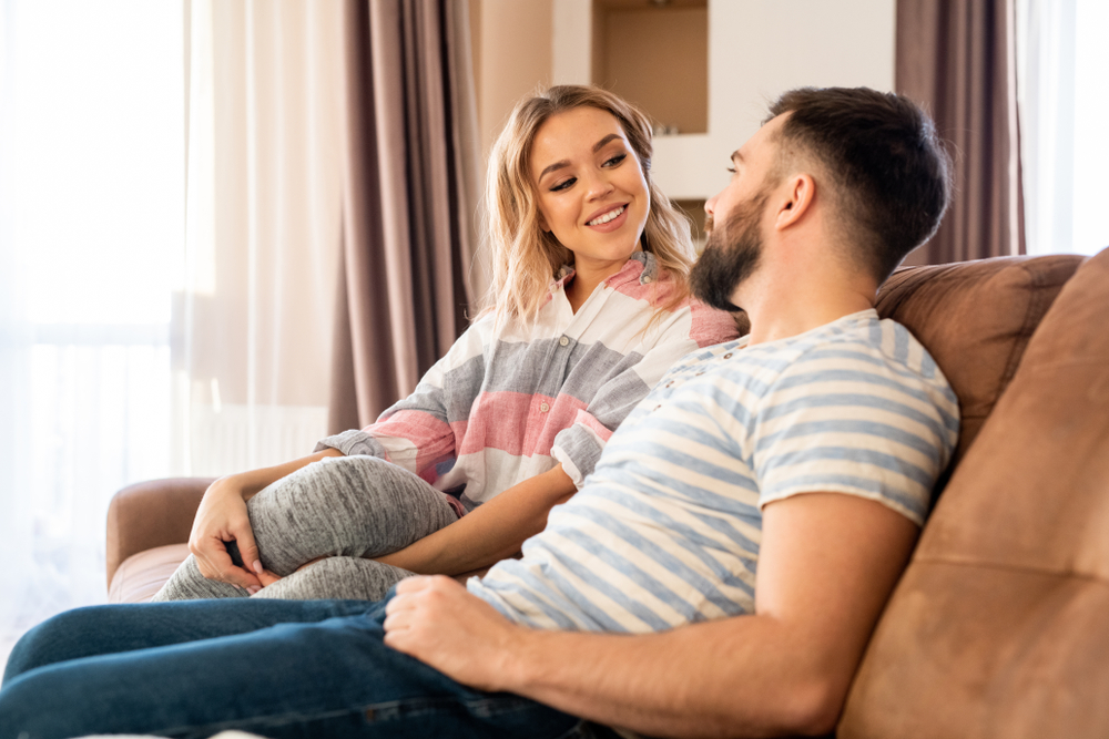 Portrait of happy couple talking to each other while relaxing sitting on sofa at home, focus on smiling beautiful woman looking at husband with love