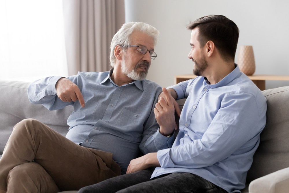 Old father wearing glasses talking with son, having pleasant conversation at home, mature dad and young man or grandson sitting on couch, discussing, sharing thoughts, family spending time together