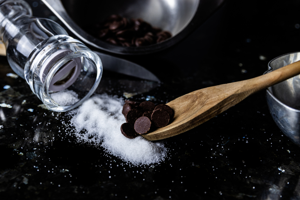 Salted dark chocolate ingredients.  Chocolate bits on a wooden spoon being added to salt that has spilled out of the salt shaker.  Health. Food trend.  Salty and sweet.