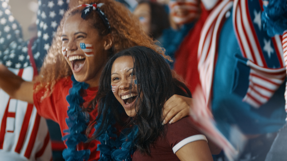 Excited fans with US flag in sports crowd celebrating on team success. Group of American soccer fans cheering in stands.