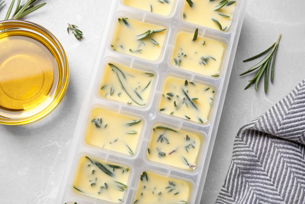 Ice cube tray with herbs frozen in oil and fresh rosemary on grey table, flat lay