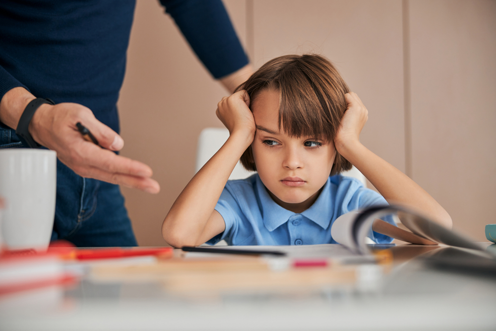 Upset Caucasian boy sitting at the table with his head leaning on the hands and looking away. Hand of his father pointing to him