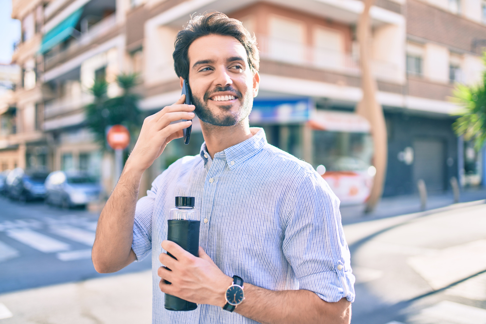 Young hispanic man holding bottle of water and talking on the smartphone at city.