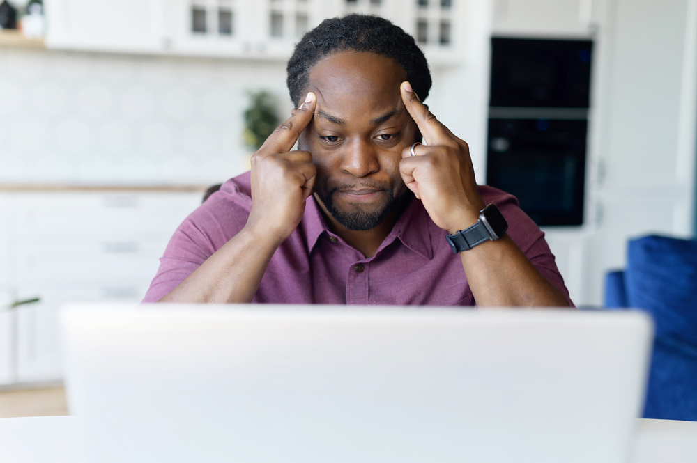 Concentrated African male freelancer using laptop for remote work at home, dark skinned man absorbed with reflections, solving difficult business tasks, staring at the screen focused