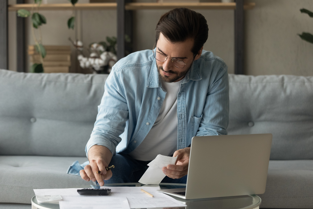 Concentrated young 30s man in eyeglasses calculating expenditures, holding paper bank bills in hands, managing monthly budget, planning investment or making online payment using computer e-banking app