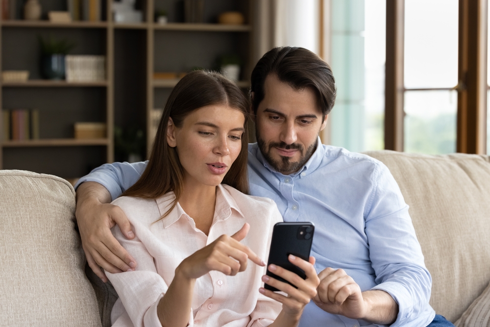 Beautiful millennial couple in love sit on sofa with smartphone, look at device screen discussing purchase, choose goods items for house using e-services at home. Modern tech, new mobile app concept
