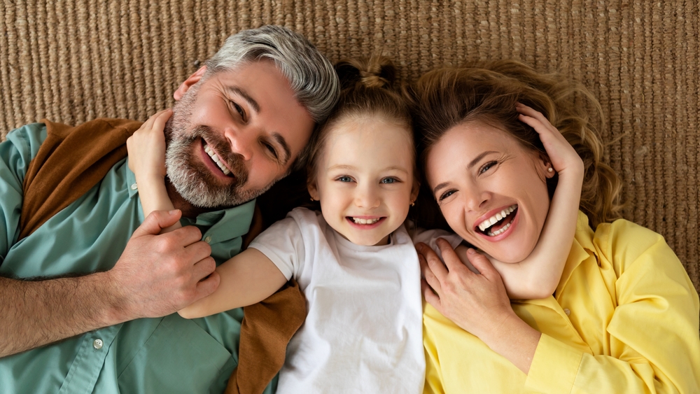 Happy Family. Joyful Parents And Daughter Embracing Lying On Floor Posing Looking At Camera At Home On Weekend. Little Daughter Hugging Mommy And Daddy. Happiness Concept. Panorama, Top View