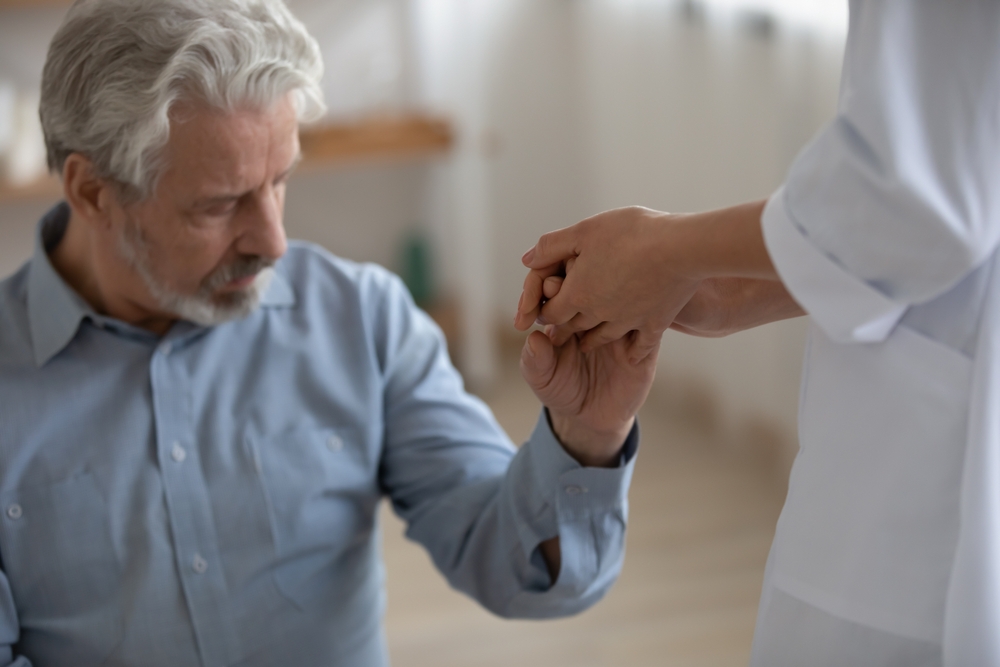 Close up nurse caregiver holding lonely mature patient hand, expressing support and care, psychological help, young woman doctor wearing white uniform comforting upset depressed senior man