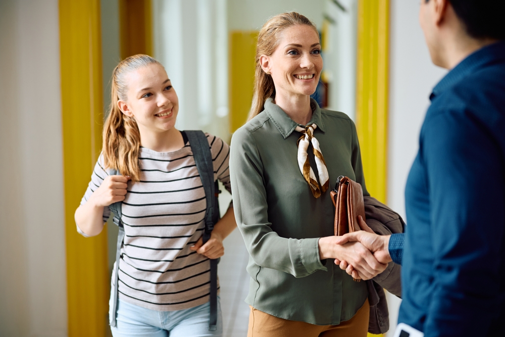 Happy mother of high school student handshaking with a principal in school hallway.