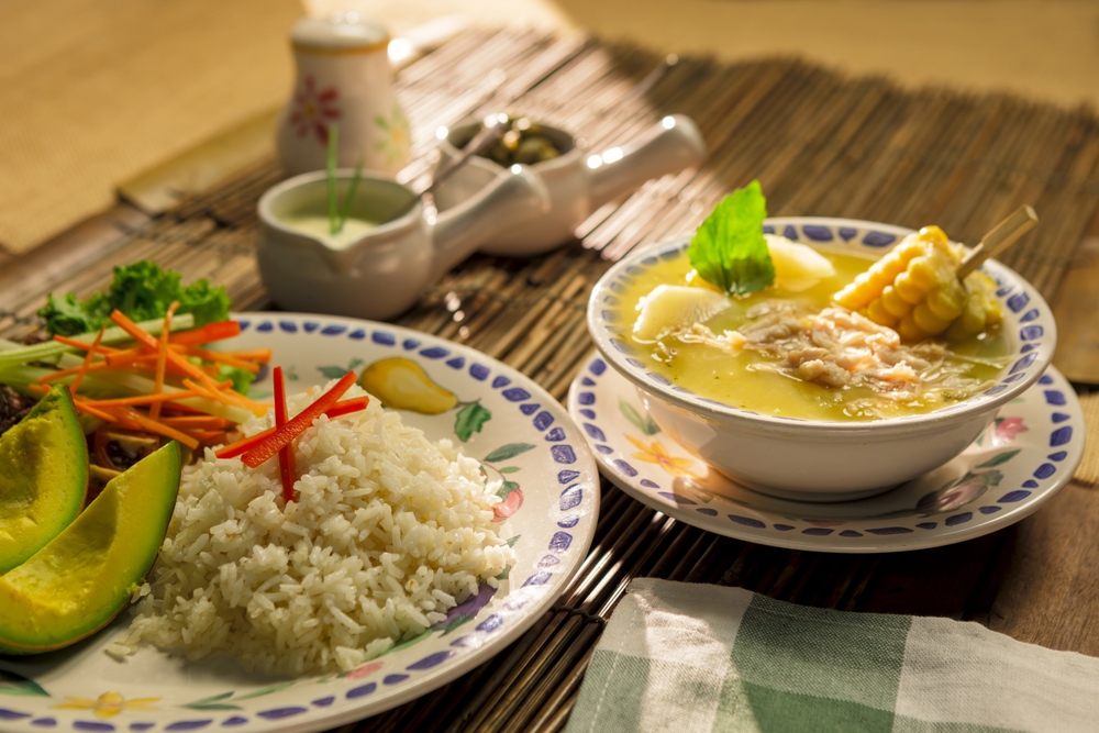 Traditional Colombian Ajiaco soup with chicken, corn, and potatoes, served with rice, avocado, cream, and capers.
