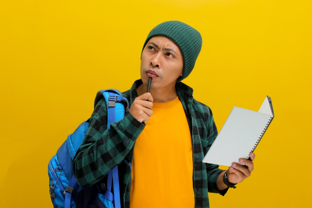 A pensive young Asian student, dressed in a beanie hat and casual shirt, carrying a backpack and holding a notebook, is engrossed in tackling a challenging assignment, deep in thought, exploring ideas