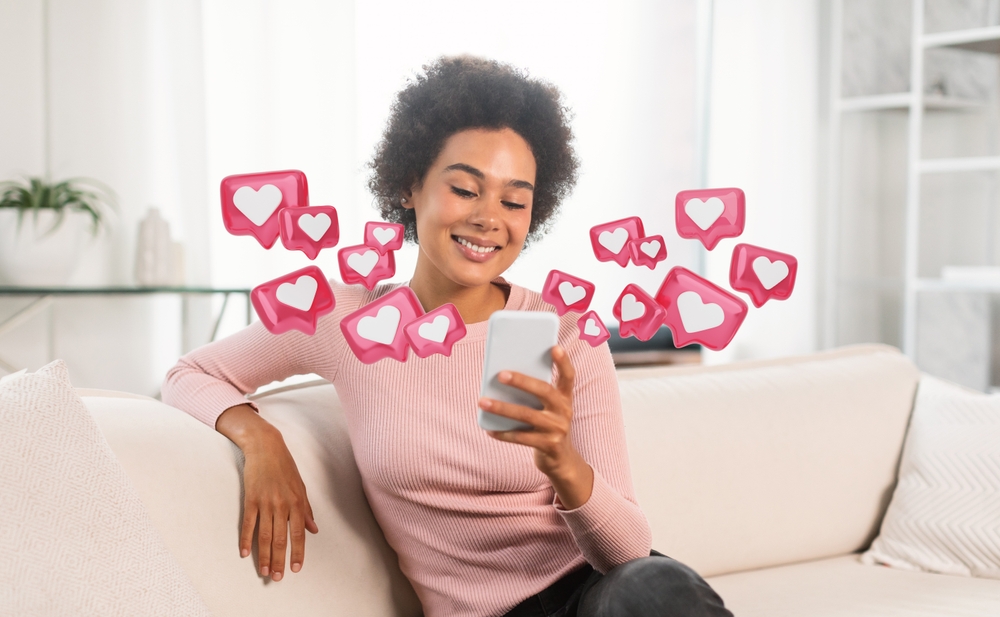 A young Black woman with curly hair is sitting on a white couch in a living room, smiling as she looks at her smartphone. She is wearing a light pink sweater and has her arm resting on the couch