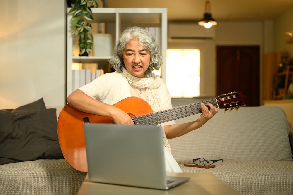 Happy elderly woman learning guitar online on laptop. Lifelong learning and modern technology for seniors concept
