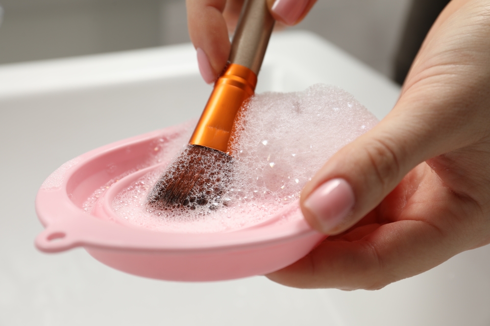 Woman washing makeup brush with soap and cleansing pad over sink, closeup