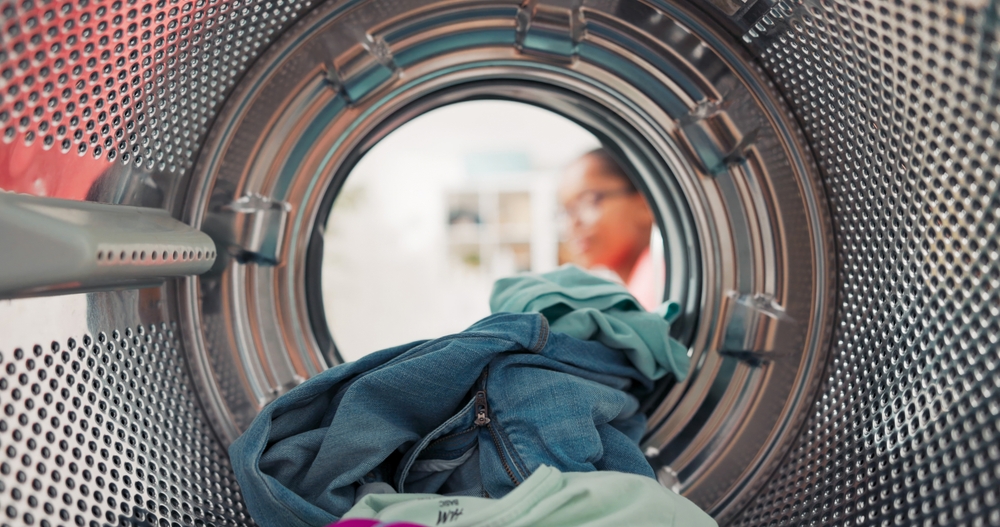 Shot from inside the washing machine drum, a woman opens the door and loads colorful clothes, including t-shirts and jeans, preparing to start a new laundry cycle.