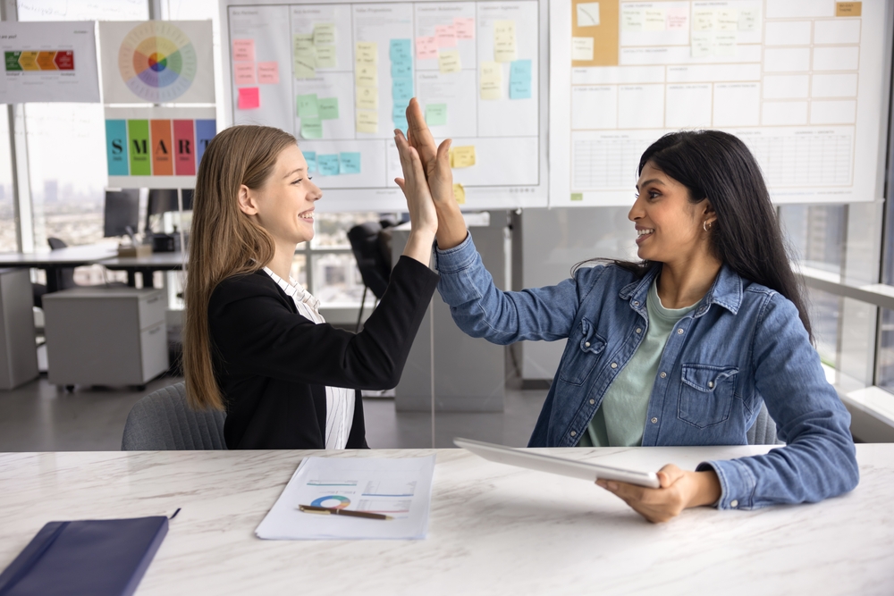 Two happy young diverse female office friends making high five gesture, enjoying successful teamwork, finding creative solution for work project, getting agreement, closing challenging task