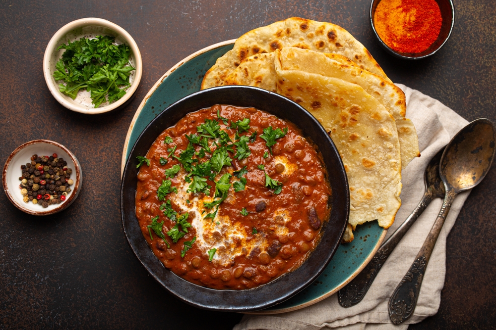 Traditional Indian Punjabi dish Dal makhani with lentils and beans in black bowl served with naan flat bread, fresh cilantro and two spoons on brown concrete rustic table top view