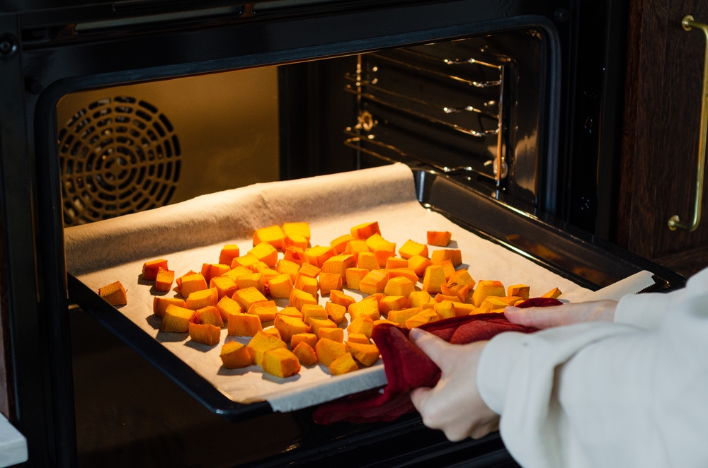 Woman removing baking tray with roasted pumpkin cubes on baking paper from oven. Preparing fresh vegetables for Thanksgiving dinner