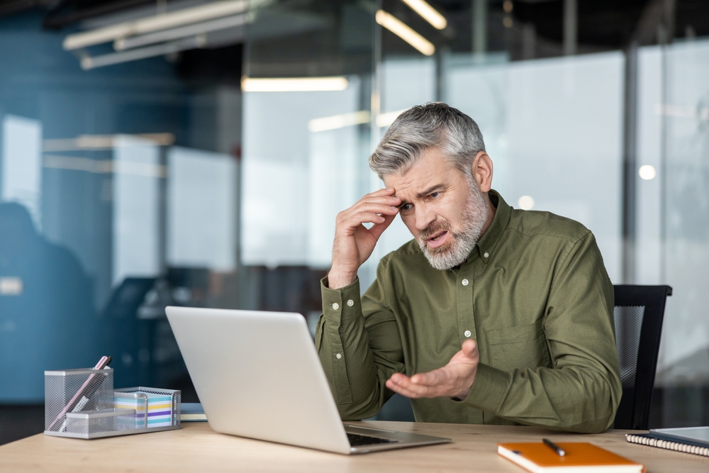Mature businessman working on a laptop in a modern office, feeling stressed, confused, and frustrated with a problem, expressing emotional difficulties and the pressure of work