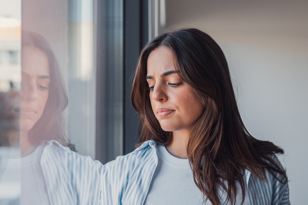 A thoughtful young woman gazes out the window with a concerned expression, captured in a casual portrait that represents uncertainty, decision making, and emotional reflection at home