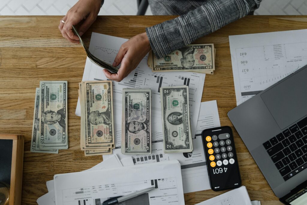 Overhead shot of hands counting cash at a wooden desk, stacks of bills arranged beside financial documents, a smartphone calculator, and a laptop.