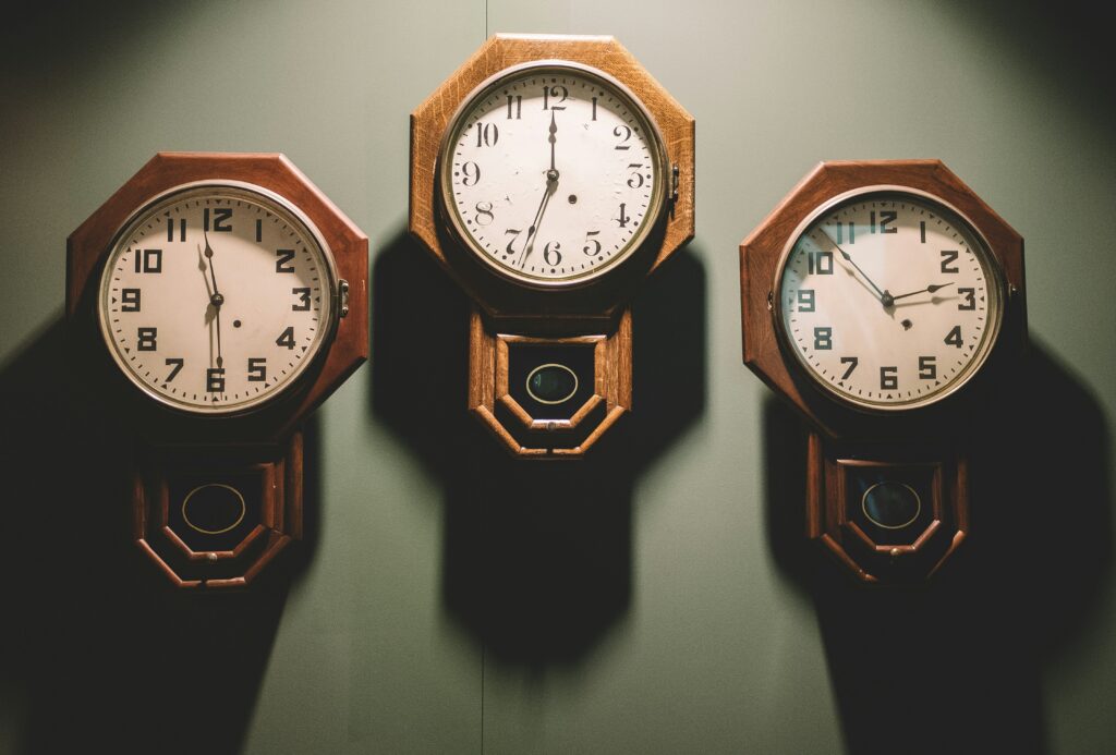Three antique wooden pendulum clocks mounted on a dark green wall, each face showing a different time, casting soft shadows across the surface.