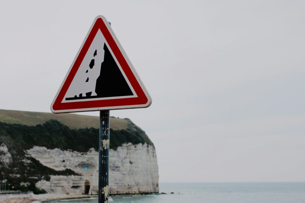 Red and white triangular falling rocks warning sign mounted on a worn metal post, with chalk-white cliffs and the sea stretching out behind it under an overcast sky.