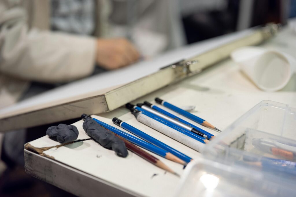A photograph of drawing supplies laid out on a white surface. Several blue mechanical pencils and lead holders sit alongside pieces of dark kneaded eraser. A hand is faintly visible in the background, resting near a drawing board.