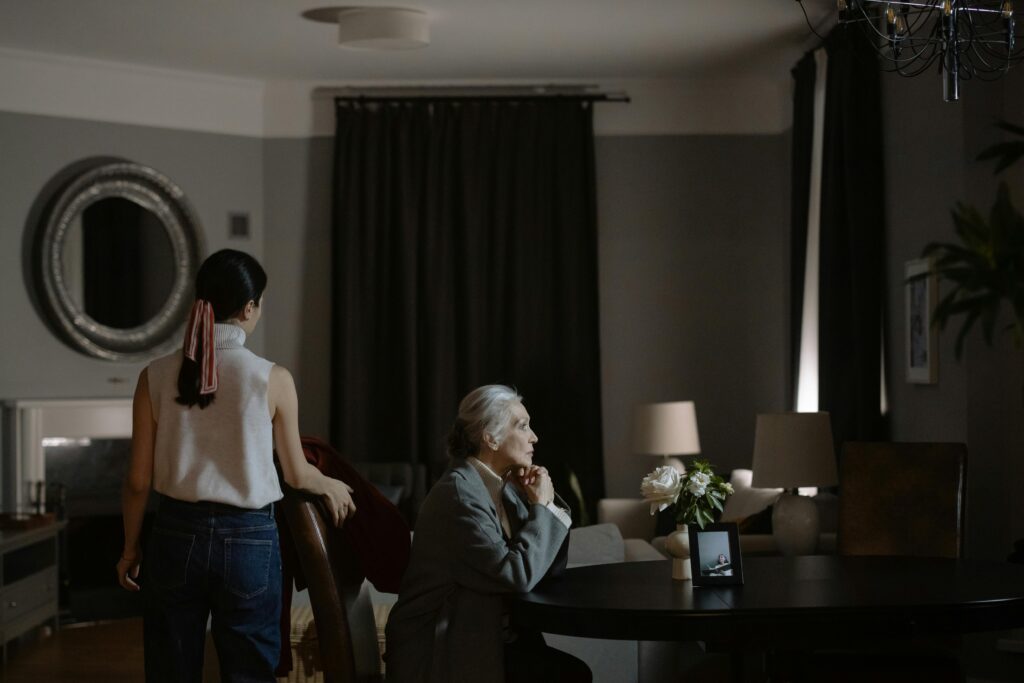 A young woman stands near a doorway watching an older woman sit alone at a dining table in a dimly lit room, suggesting emotional distance.