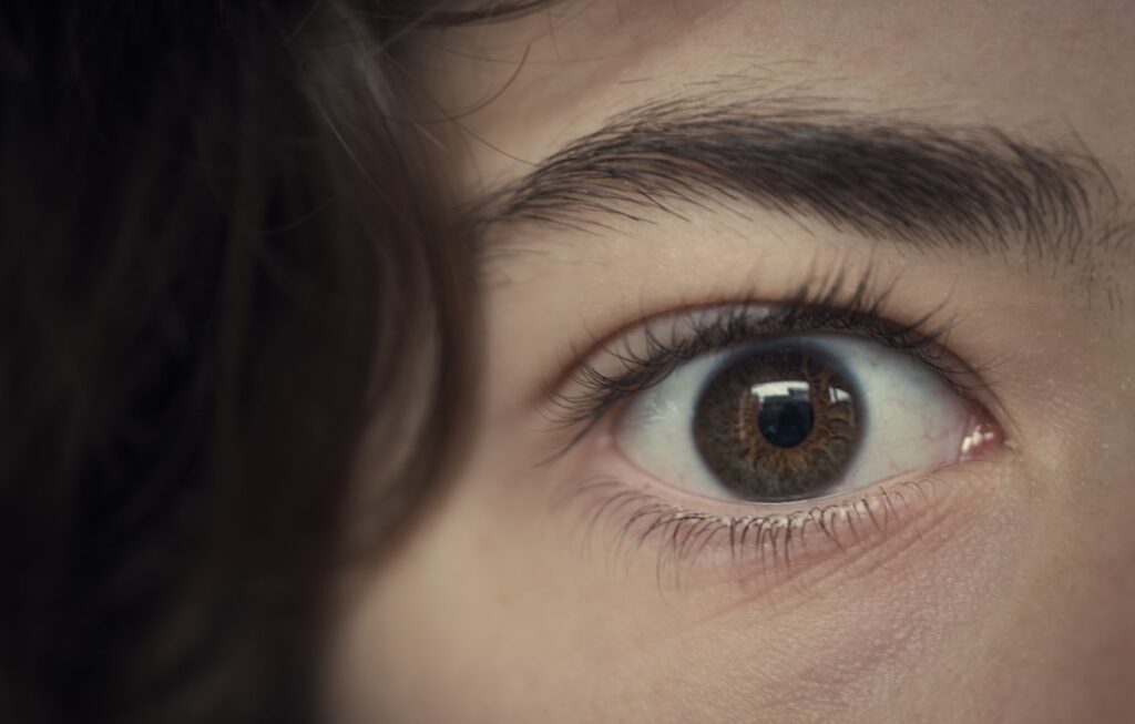 A close-up photograph of a brown eye with flecks of hazel in the iris. Dark hair falls across the forehead, and the eyebrow and surrounding skin are slightly out of focus.