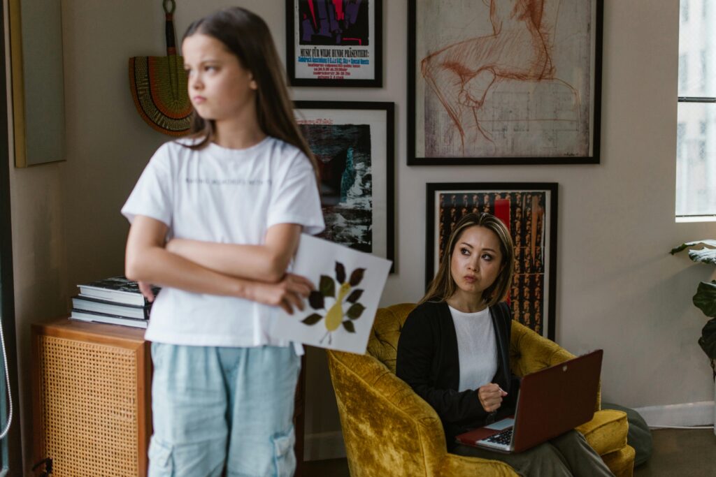 A young girl stands with arms crossed and back turned, holding a drawing, while a woman sits in a yellow armchair with a laptop, looking over at her with concern.