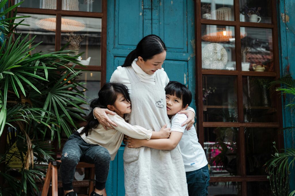 A mother stands in front of a blue doorway hugging her young daughter and son close on either side of her, surrounded by green plants.