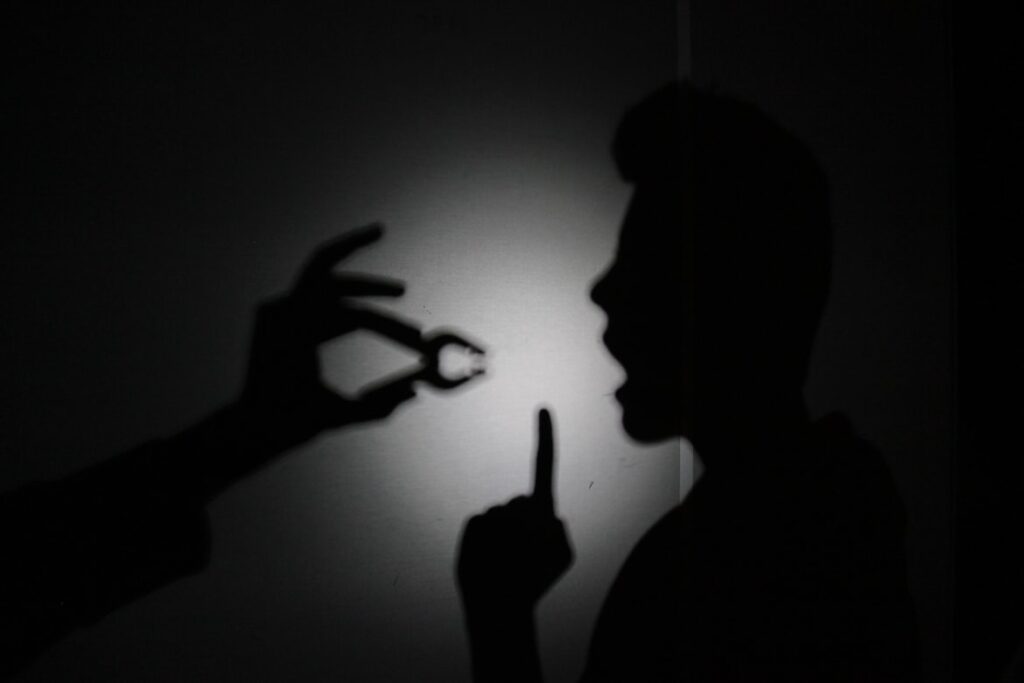 A dark silhouette of a person on a wall, with a shadow hand pinching their lips closed while the person holds a finger to their mouth in a shushing gesture.