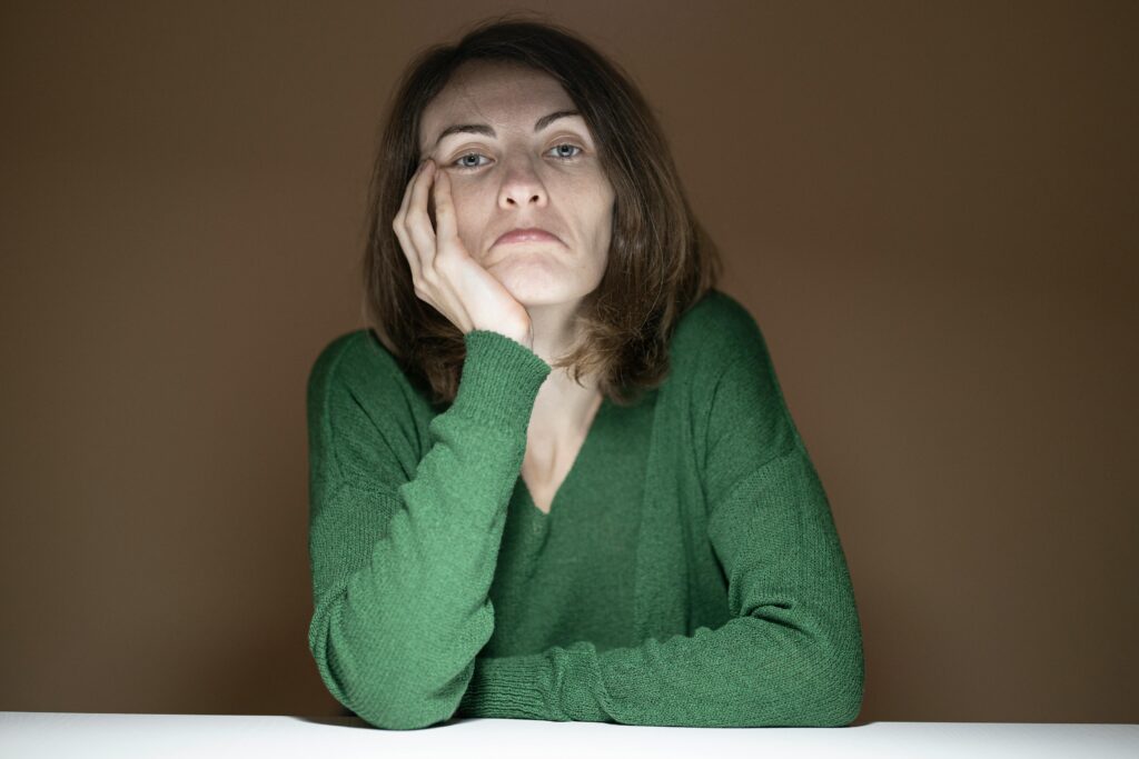 A woman in a green sweater rests her cheek on her hand at a table, staring directly at the camera with a tired, flat expression.