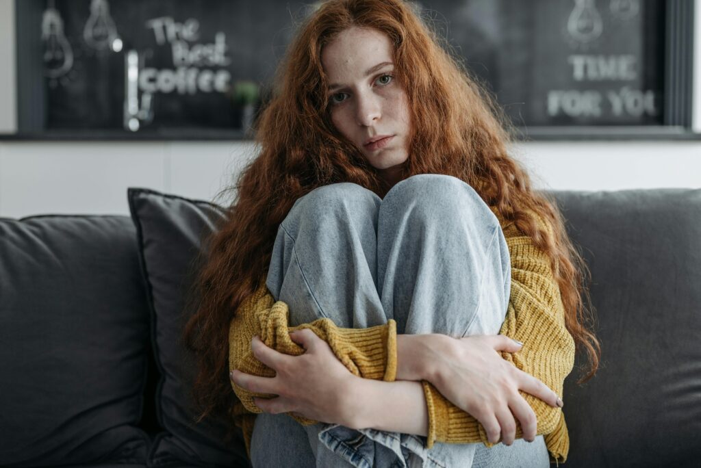 A young woman with long red hair sits curled up on a grey couch, hugging her knees to her chest in a yellow sweater, staring at the camera with a sad expression.