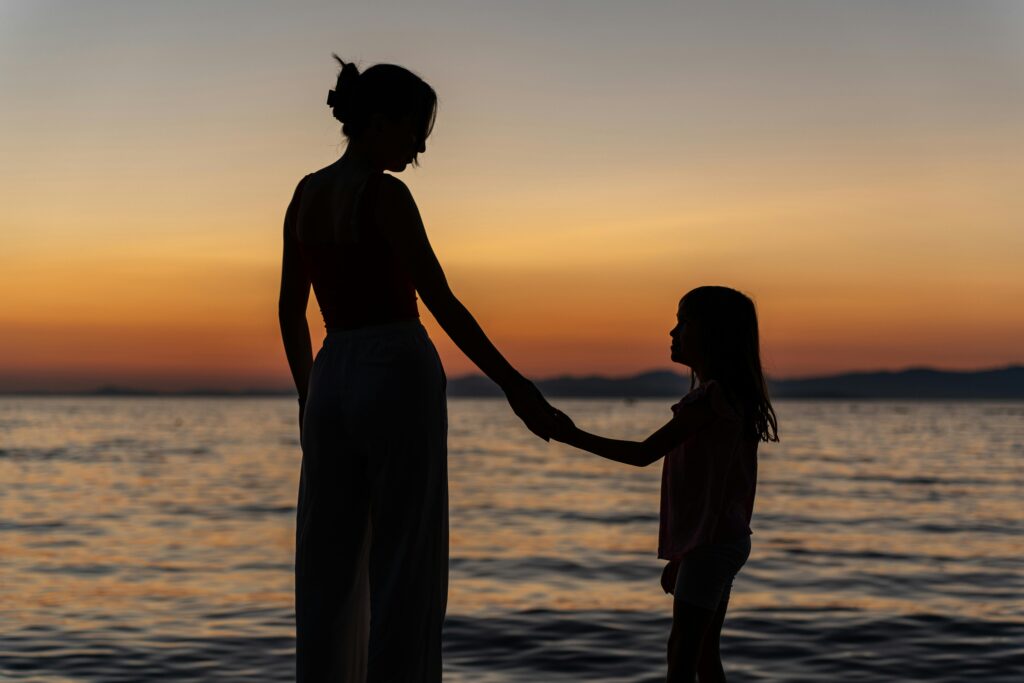 The silhouettes of a woman and a young girl stand holding hands by the ocean at sunset, facing each other against a glowing orange sky.