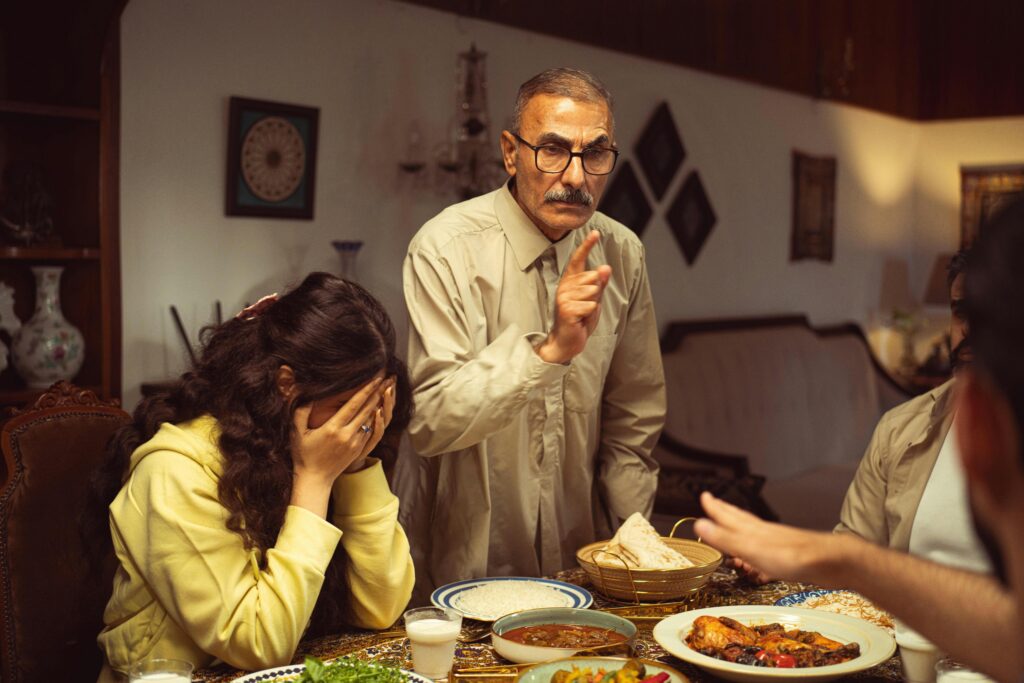 An older man with glasses and a moustache stands at a dinner table, pointing and scolding a young woman who covers her face with her hands in distress.