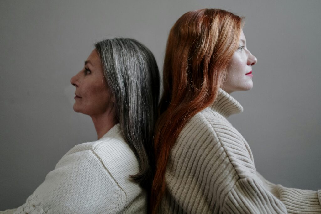Two women in cream knitted sweaters stand back to back, one with long grey hair and the other with long red hair, both looking in opposite directions against a plain wall.