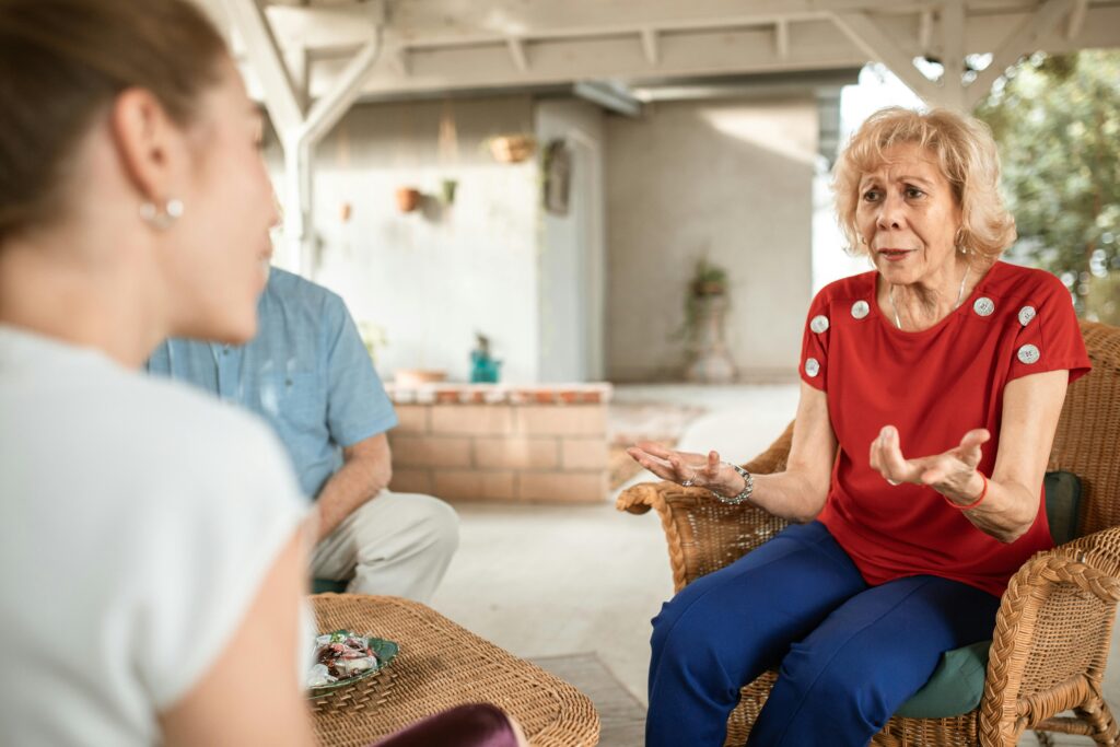 An older woman in a red top sits in a wicker chair, gesturing with both hands as she speaks earnestly to a younger woman seated across from her.