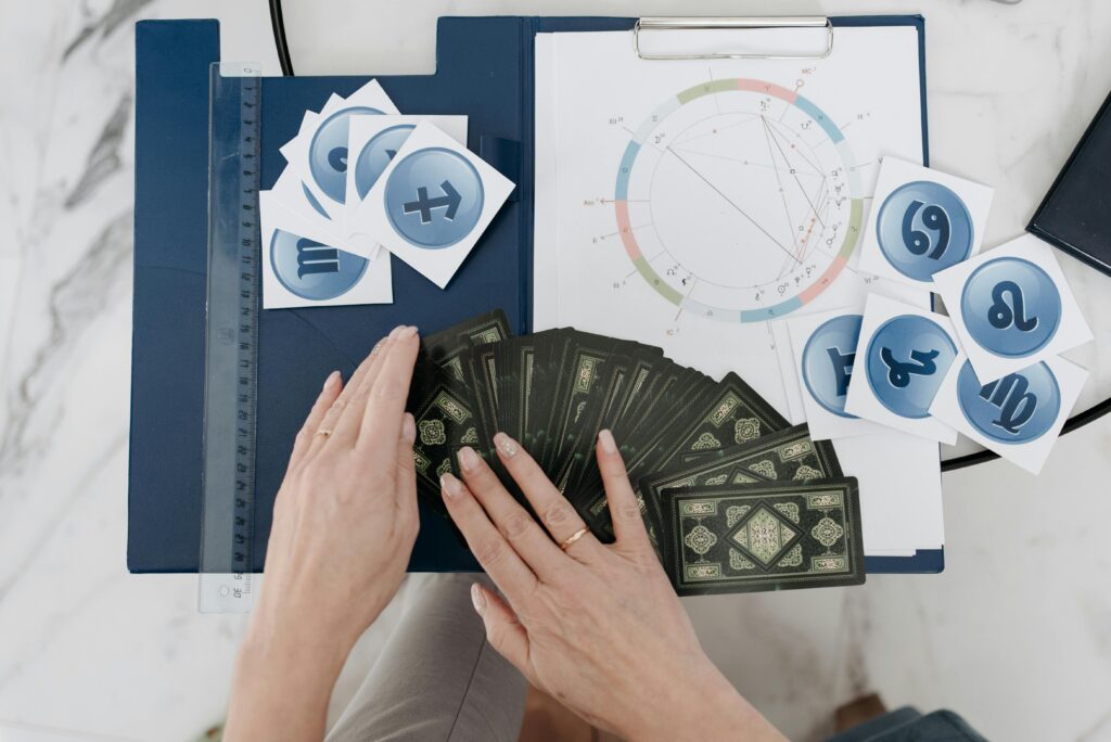 Overhead view of a person shuffling tarot cards on a navy mat, with zodiac symbol tiles and an astrological birth chart spread across a marble surface.